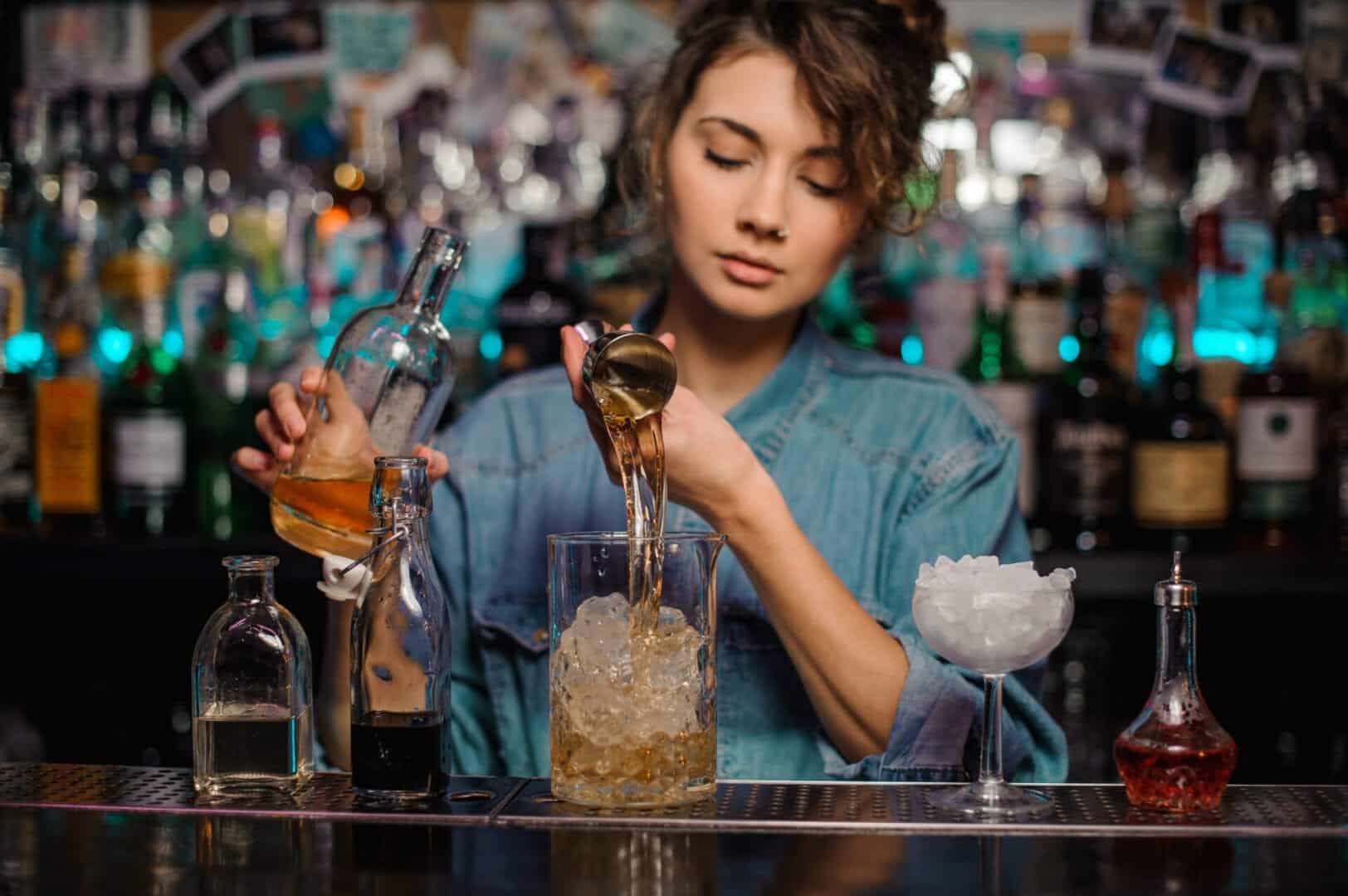 Bartender girl pouring to the measuring glass cup with ice cubes an alcoholic drink from jigger on the bar counter on the blurred background